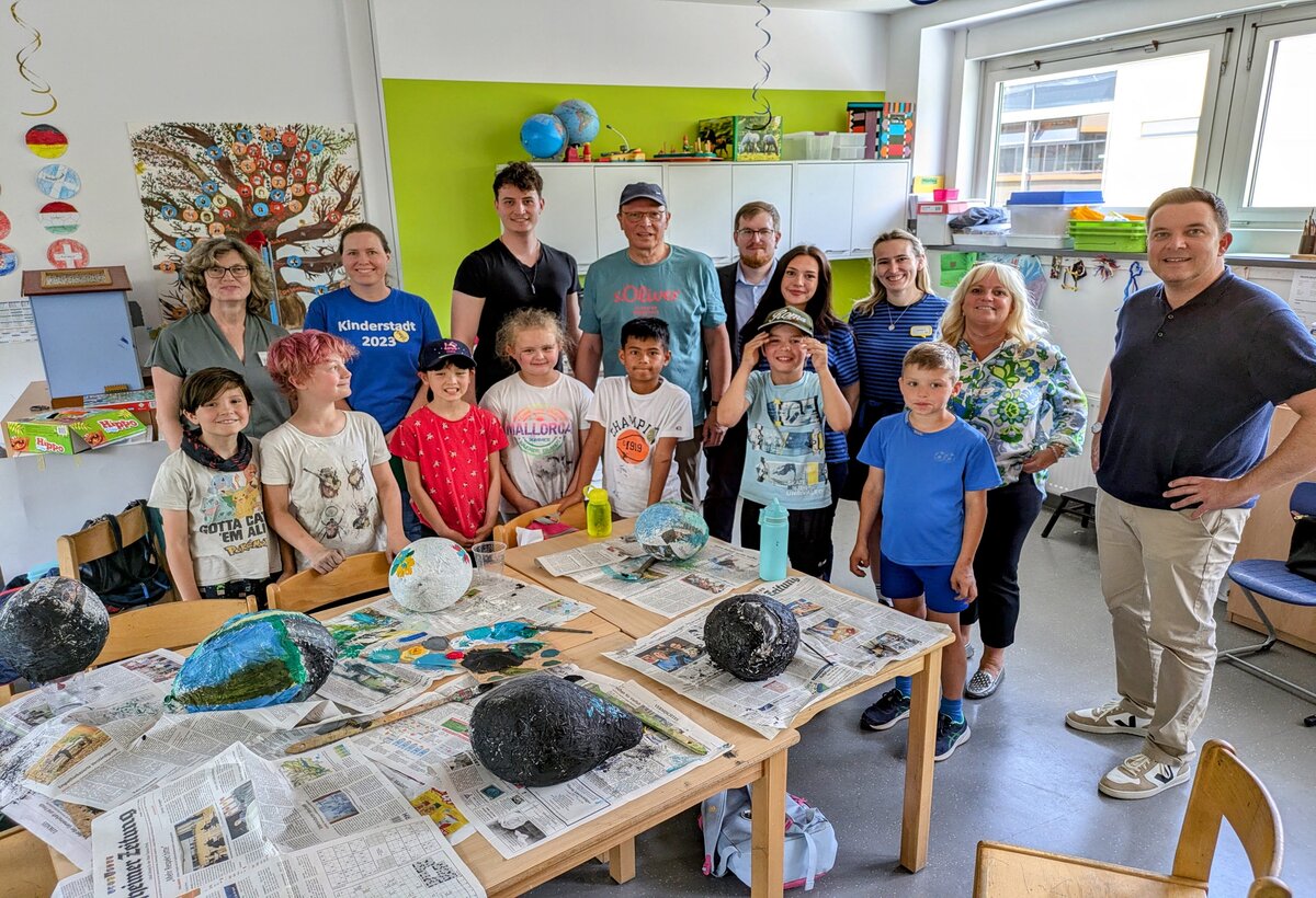 Bürgermeister Christian Vogt (rechts), Fachbereichsleiterin Susanne Schindler (links), Leiter der Ferienspiele Bernd Schick (Mitte mit Kappe) und die Sponsoren Susanne Ebert von der Taunus Sparkasse (Zweite von rechts), Kevin Bornath von der Mainova (hinten mit Brille) sowie Annabell Großmann und Hannah Zehnder von Ikea (in gestreiften T-Shirts) mit einer Gruppe der Ferienspiele in einem der Bastelräume in der Heiligenstockschule.