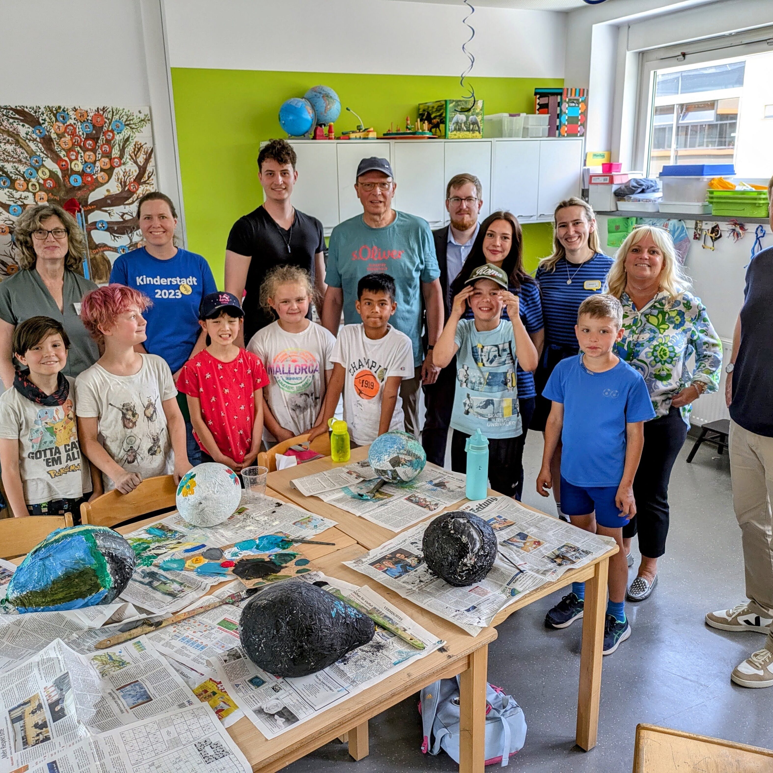 Bürgermeister Christian Vogt (rechts), Fachbereichsleiterin Susanne Schindler (links), Leiter der Ferienspiele Bernd Schick (Mitte mit Kappe) und die Sponsoren Susanne Ebert von der Taunus Sparkasse (Zweite von rechts), Kevin Bornath von der Mainova (hinten mit Brille) sowie Annabell Großmann und Hannah Zehnder von Ikea (in gestreiften T-Shirts) mit einer Gruppe der Ferienspiele in einem der Bastelräume in der Heiligenstockschule.