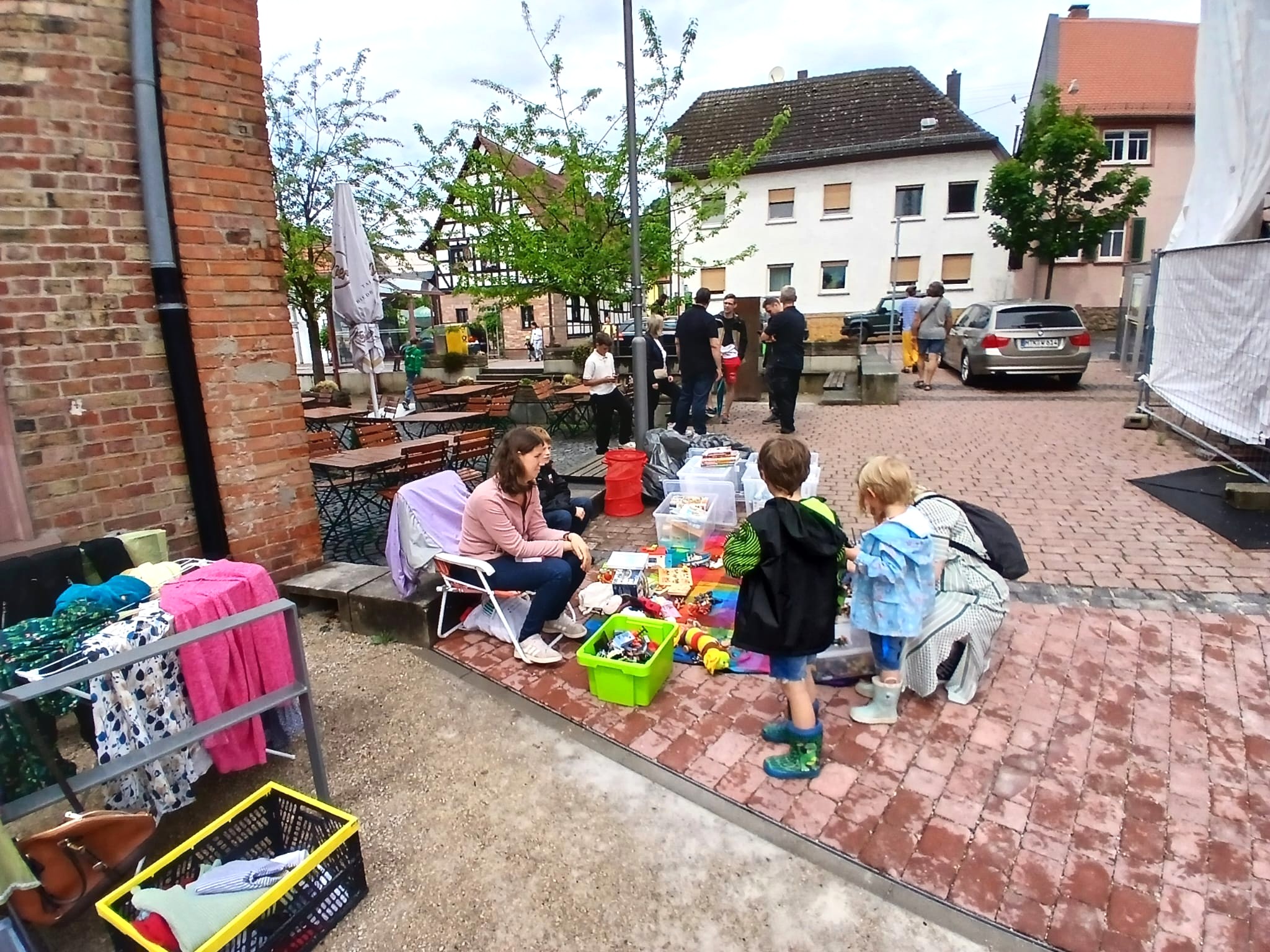 Dorfflohmarkt Diedenbergen, Stand an dem eine Mutter mit Kind Sachen verkauft und eine junge Familie gerade die Angebote durchsucht